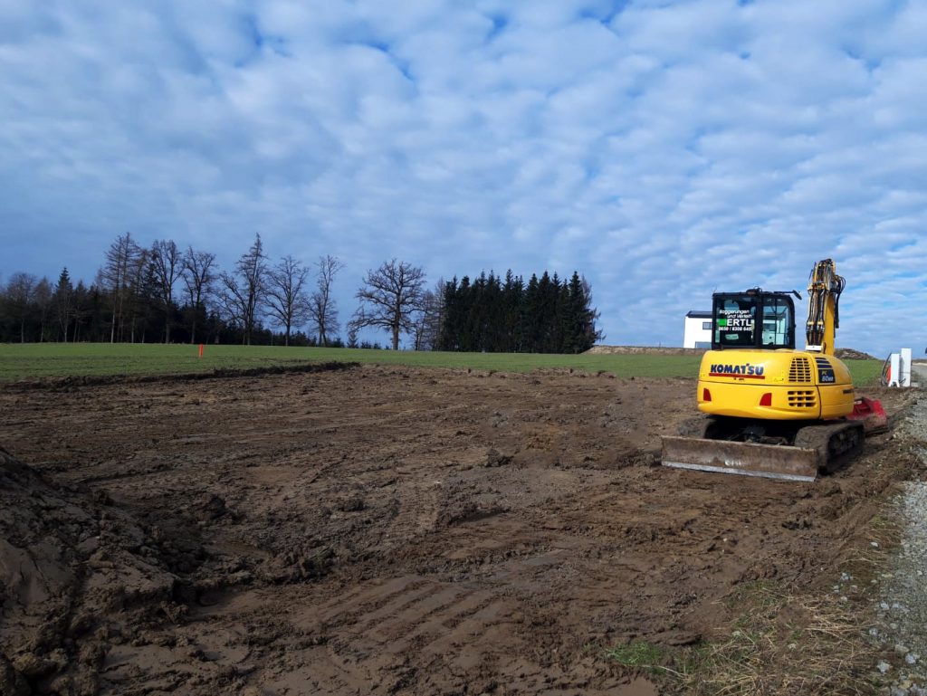 Unser Bagger im Einsatz. Man sieht die Baggerungen und Erdarbeiten. Im Hintergrud ein bewölkter Himmel und einige Bäume die hinter dem Feld stehen.